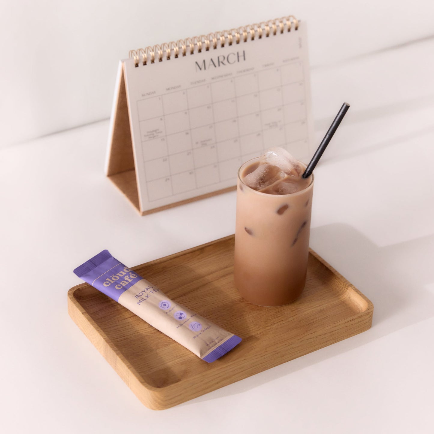 A glass of iced Cloud Cafe Royal Milk Tea with a straw sits on a wooden tray next to a instant milk tea packet and a desk calendar displaying the month of March.