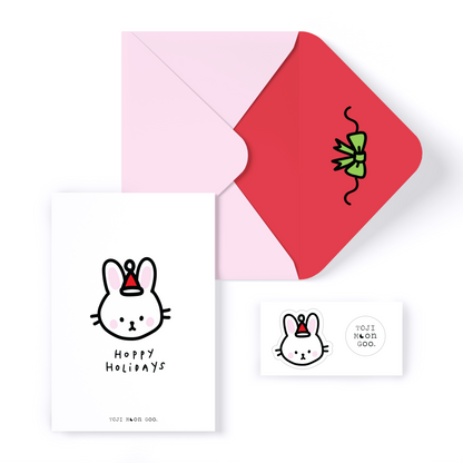 White minimal card with bunny head doodle on front, reading "hoppy holidays". Bunny is wearing a small red Santa hat. Envelope is light pink with red inner color and green doodle bow. 2 stickers shown: bunny head with santa hat and TOJI MOON GOO white logo circle sticker.