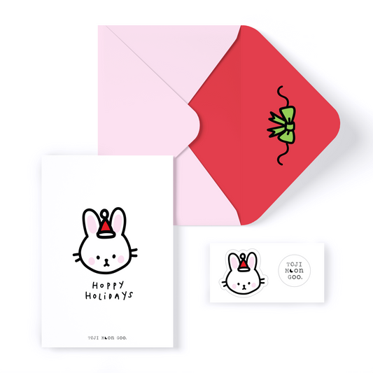 White minimal card with bunny head doodle on front, reading "hoppy holidays". Bunny is wearing a small red Santa hat. Envelope is light pink with red inner color and green doodle bow. 2 stickers shown: bunny head with santa hat and TOJI MOON GOO white logo circle sticker.